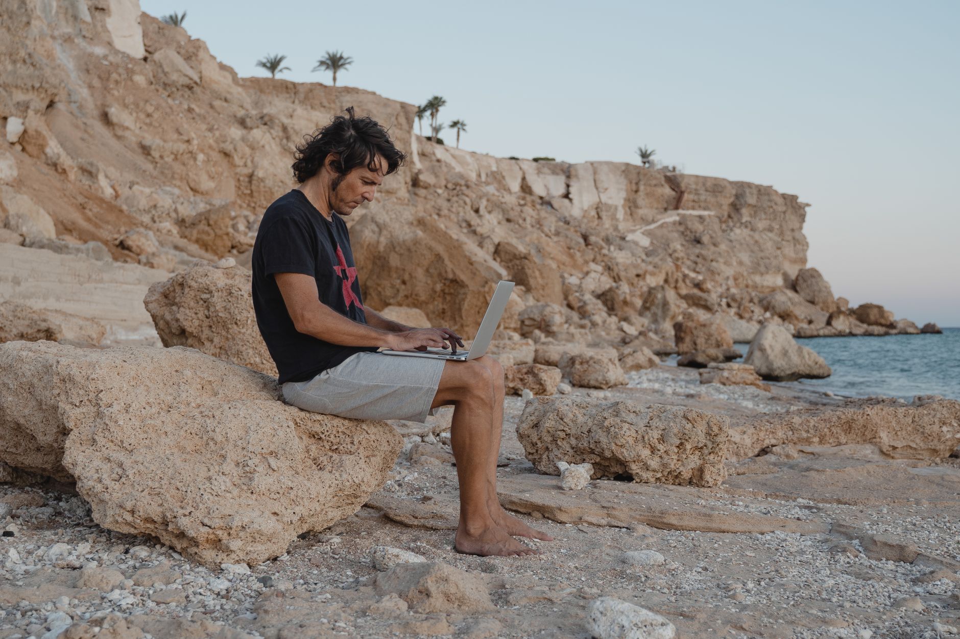 Homme assis sur des rochers avec un ordinateur portable au bord de la mer, travaillant à distance au coucher du soleil.