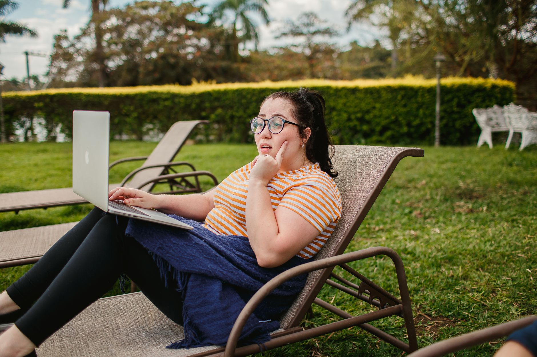 Femme se relaxant sur une chaise longue avec un ordinateur portable dans un jardin verdoyant, un jour ensoleillé.