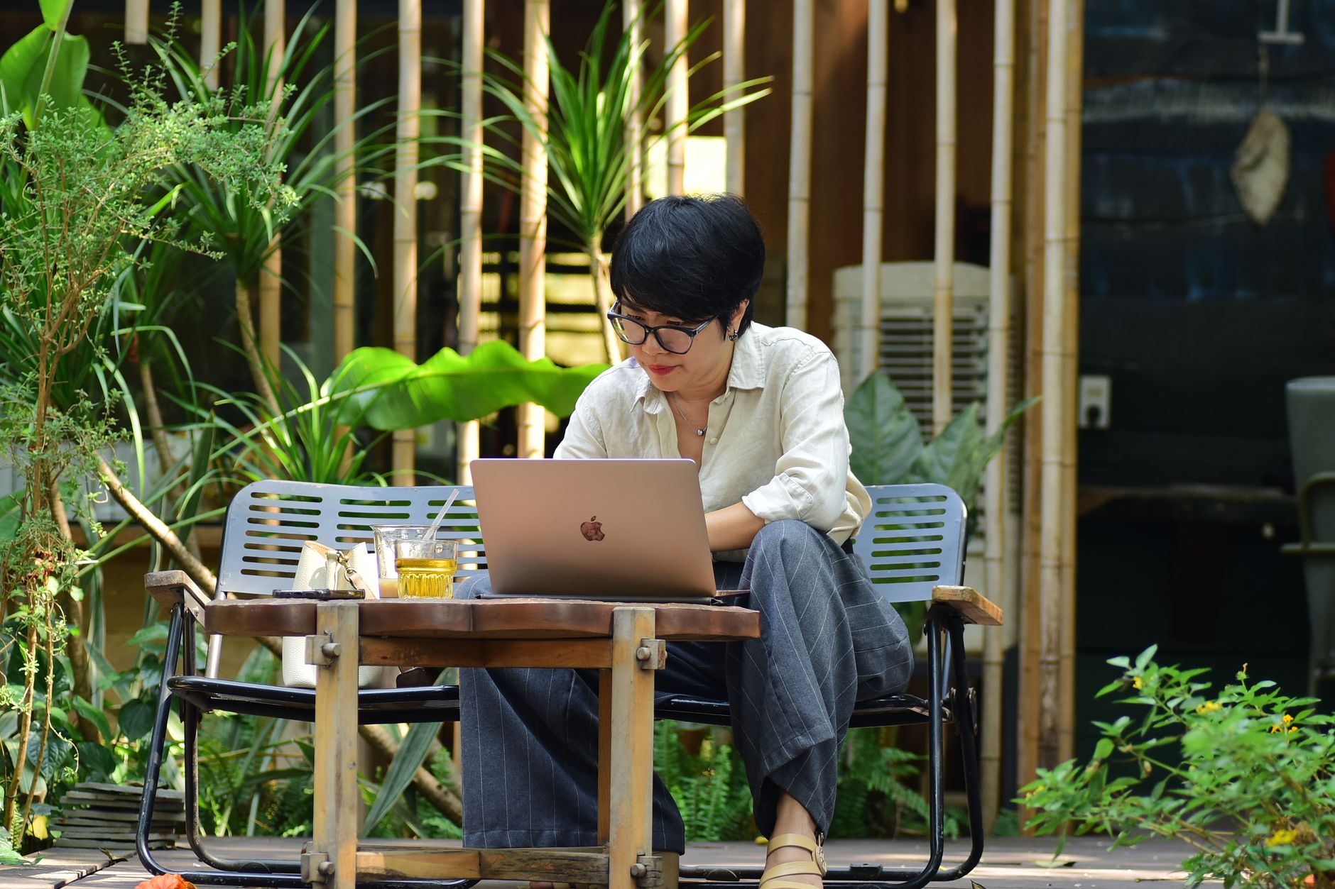 Une femme concentrée sur son ordinateur portable, assise dans un café extérieur verdoyant.